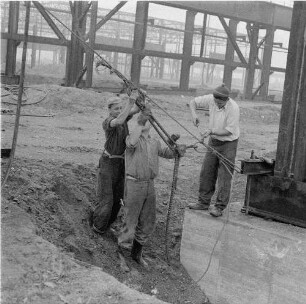 Zeithain. Baustelle Rohrwerk III des VEB Stahl- und Walzwerk Riesa. Produktionshalle (Stahlskelettbau) im Rohbau. Drei Bauarbeiter beim Spannen einer Seilarmatur