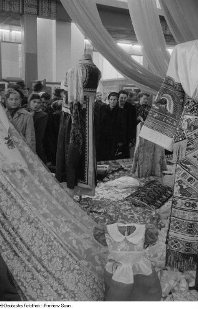 Besucher vor einem Messestand mit Stoffen und traditionellen Kleidern auf der Ausstellungsfläche der Tschechoslowakei