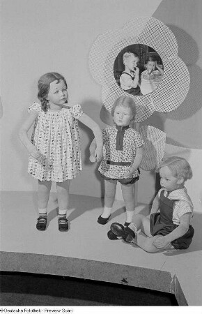 Kinderbekleidung präsentiert an Schaufensterpuppen auf der Leipziger Herbstmesse 1954 dekoriert mit einer Fotografie zweier speisender Kinder