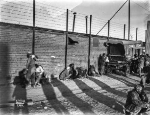Straßenszene : Einige Personen sitzen vor einer Mauer. Rechts ein Kutschenwagen der Firma Evans & Brown.
