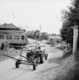 Bahnstrecke zwischen Hamburg und Lübeck: Bahnübergang an Schrankenposten 70 mit geschlossenen Schranken: davor landwirtschaftliches Fuhrwerk mit Anhänger: links fahrender Personenzug: dahinter Schrankenwärterhäuschen: hinten rechts und Mitte Knick mit Büschen: dahinter Weide