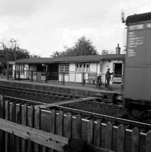 Bahnhofsgebäude: Bahnsteig: Gleise: Bahnangestellter: rechts Teil eines Güterwagens in Bewegung: im Vordergrund Lattenzaun