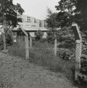 Blick über den Maschendrahtzaun mit Stacheldraht (ehem. Grenze des Geländes der Staatssicherheit) des Gebäudekomplexes Nr. 112 - 122 gegen eine Baracke, ein Nebengebäude und die östlich angrenzende 5-geschossige Wohnbebauung