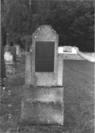 Grabstein 71 (Index: 99609707) Material: Kunststein?, Platte Granit? Maße HxBxT: 186x71x16 cm Beschreibung: Stele, mehrfach geschwungener eingezogener Segmentbogenabschluß, Seitenwangen, rechteckige Schriftplatte eingelassen, getreppter Sockel: Grabstein 71
