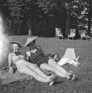 Zwei Frauen in Bademode und einem Strohhut liegen auf einer Wiese