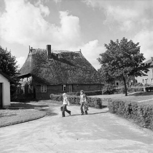 Alte Dorfstraße: Bauernhaus mit Reetdach: rechts davor Grünanlage mit Hecke und Baum: vorn zwei Schulkinder