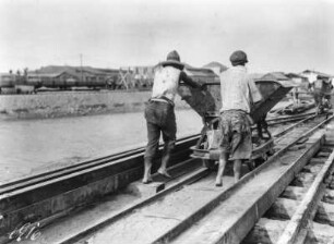 Männer schieben eine Lore : Zwei Männer schieben eine Lore am Hafen entlang