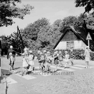 Schulstraße: Fußgängerüberweg mit Zebrastreifen und Hinweisschild: Schülerlotsen leiten Schulkinder der Schlossschule mit Schirmmützen und Rantzen über die Straße: hinten Haus mit Straßenschild an Giebel, Hecke, Strommast, Bäume