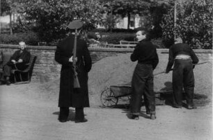 Strafvollzug. Strafgefangene bei Außenarbeiten. Sie werden von einem bewaffneten Justizvollzugsbeamten bewacht