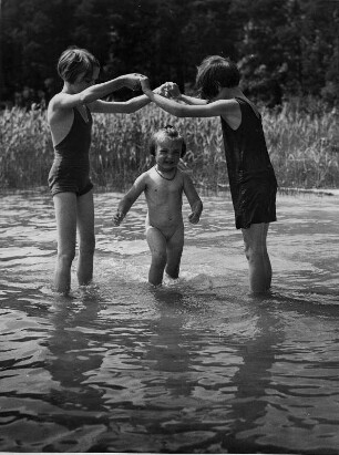 An einem Gewässer. Drei Mädchen spielen im Wasser. Sie tanzen Ringelreihen.