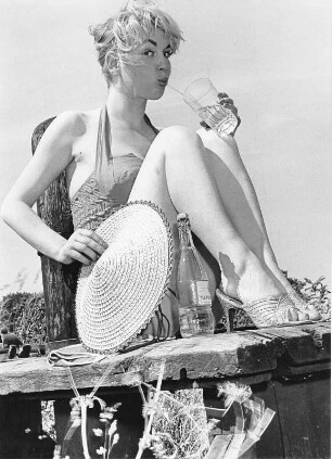 Portraitaufnahme des Fotomodels Frl. Haddorp. Sie sitzt auf einem Steg und trinkt. Aufgenommen 1957