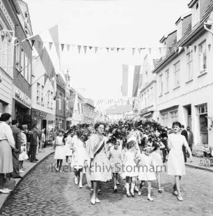 Kindervogelschießen: Umzug: Hindenburgstraße: Mädchen mit Blumenbögen: an den Straßenrändern Zuschauer: über Straße Wimpelgirlanden, an Fassaden Fahnenschmuck: an linker Straßenseite Commerzbank, Friseursalon Lübcke, hinten Gaststätte "Bierquelle": an rechter Straßenseite hinten Schuhgeschäft Paul Jürgens: im Hintergrund Hagenstraße, 2. Juli 1963