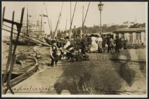 Personen warten an Anlegestelle des Bosporus auf das Eintreffen einer Fähre, fotografiert von einer Brücke in Istanbul, im Hintergrund ein großes Segelschiff und Blick auf den gegenüberliegenden Stadtteil. Beginn des Baus der Bagdadbahn in der Türkei