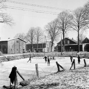 Dorfteich mit Schlittschuhläufern: dahinter Heilsaustraße: im Hintergrund Bauernhäuser und Bäume: vorn Zaun: links Mantel über Zaunpfahl und Schultaschen: Winter