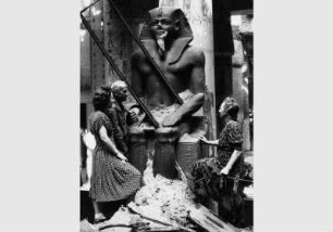 Restaurator Rudolf Kuhn mit zwei Frauen an der Kolossalstatue Sesostris II im Ägyptischen Hof im Neuen Museum