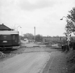 Bahnstrecke zwischen Hamburg und Lübeck: Bahnübergang an Schrankenposten 70 mit geschlossenen Schranken und Andreaskreuz: links Ende eines durchfahrenden Zuges: hinten links Dach des Postens: im Hintergrund Arbeiter mit Schubkarre: dahinter Pkws