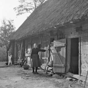 Strohgedecktes Blockhaus in Burg-Kauper. Frau Nowick in Spreewälder Alltagstracht vor ihrem Gehöft. : Ze słomu skryte kładźity twar statoka w Kupaŕskich Bórkowach. Knjeni Nowickowa we wšědnej Błótowskej drasće před swojim statokom.