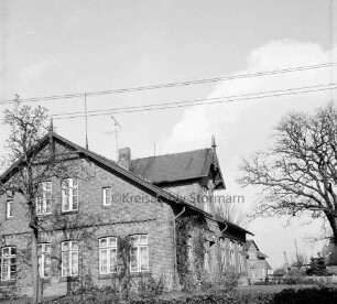 Volksschule mit Schulhof: vorn Hecke: rechts und links Bäume: hinten rechts Haus