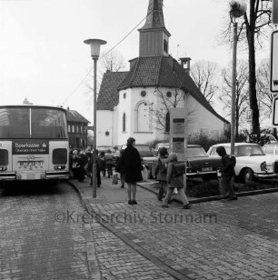 Paul-von-Schoenaich-Straße: Schulbus an Haltestelle: Schüler mit Schulranzen: Straßenlampe, Parkplatz, parkende Autos, Mast mit Scheinwerfer: hinten Matthias-Claudius-Kirche