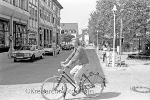 Hagenstraße: verkehrsberuhigter Bereich: links Wohn- und Geschäftshäuser: davor parkende Pkws: rechts Bäume und Anlage vor Kreissparkasse Stormarn mit Fußgängern: vorn Fahrradfahrer: im Hintergrund Gebäude