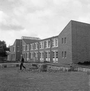 Bargteheide: Kreisgymnasium II [später Gymnasium Eckhorst]: Eckhorst: vor Fertigstellung: Herrichtung der Außenanlagen: Arbeiter beim Pflastern des Halbrunds mit Treppen und Anpflanzungen, Schubkarre: links Lüftungsrohr