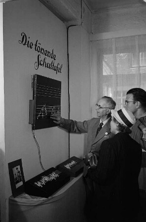 Drei Personen begutachten im Zentral-Messepalast auf der Leipziger Herbstmesse 1955 "Die tönende Schultafel" der Firma Doremi