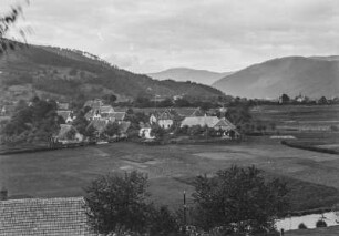 Dorf : Ein Dorf umgeben von Ackerland in einem Hochtal. Im Hintergrund Hügelketten mit Bergwald, im Vordergrund ein Bach