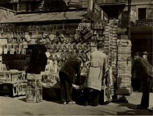 Barcelona. Spanien. Einer von vielen großen Zeitungsständen auf der Rambla del Centro, der Zentralstraße der Stadt. Ein Verkäufer sortiert die Auslagen. Ein Kunde sucht sich Lesestoff aus