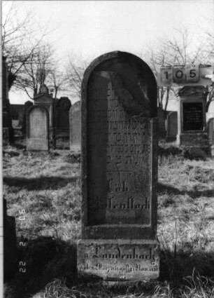 Grabstein 105 (Index: 100116415) Material: Sandstein Zustand: rechter oberer Teil der Stele abgebrochen mit Textverlust Maße HxBxT: 165x56x25 cm Beschreibung: aufgesockelte Stele mit Rundbogenabschluß, eingetieftem Schriftfeld und Schriftfeld am Sockel: Grabstein 105