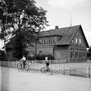 Volksschule: links davor Garten mit Baum: rechts Schulhof: vorn Zaun: davor zwei Kinder auf Fahrrädern