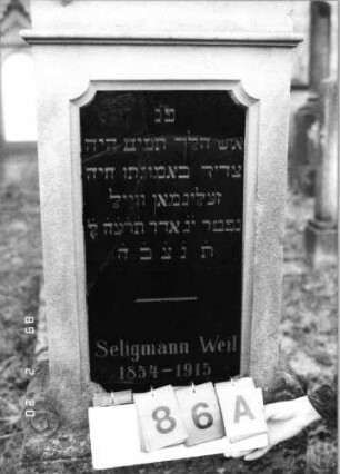 Grabstein 86 (Index: 99798547) Material: Sandstein, Schriftplatte Granit Zustand: gut, Schriftplatte gesprungen Maße HxBxT: 176x46x25 cm Beschreibung: Stele mit Volutenaufsatz, Schriftplatte mit geradem Aufsatz, Sockel: Grabstein 86A