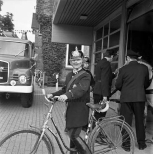 Tremsbüttel: Amtsfeuerwehrfest und 90-jähriges Jubiläum der Freiwilligen Feuerwehr Tremsbüttel: Jungfeuerwehrmann Olaf Tödt in historischer Uniform mit Feuerhorn und Fahrrad: im Hintergrund rechts Feuerwehrgerätehaus, im Eingang zwei Männer mit Pferdegespann: links Löschfahrzeug