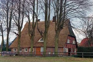 Kamp 12: Fachwerkhaus mit Reetdach: Traufseite, von links ehemaliger Wirtschaftstrakt, Sprung, Tür, Wohntrakt: Giebelseite Wohntrakt mit Tür, Sprossenfenster: Fledermausgaube, Giebelzier Pferdeköpfe: davor Baumreihe, Holzzaun