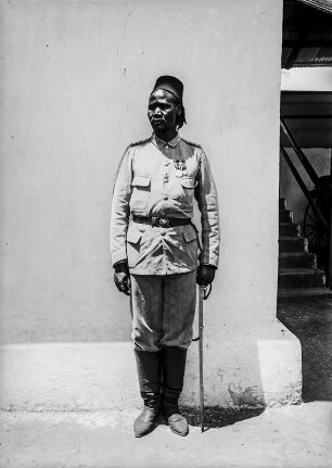 Deutscher Kolonialsoldat : Portrait eines deutschen Kolonialsoldaten, ausgestattet mit einem Säbel, vor einer weißen Mauer stehend