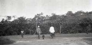 Zwei Männer in Tropenkleidung mit zwei Hunden auf einem Weg vor einer Siedlung auf einer Anhöhe im Hintergrund