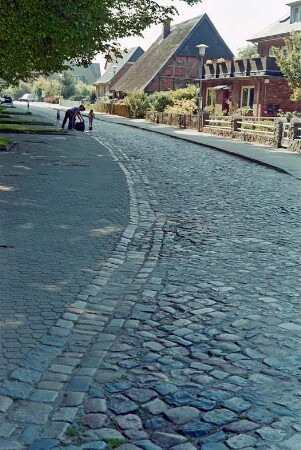 bei Kirche: Straße mit Kopfsteinpflaster, Rinnstein: rechts Häuser, links Parkfläche