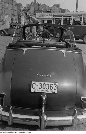 Ein geparkter Chevrolet mit amerikanischem Autokennzeichen, im Hintergrund ein Omnibus, ein Imbiss und Gebäuderuinen
