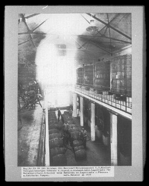 Weinbrennerei C. T. Hünlich, Wilthen. Innenansicht, zweigeschossiges Gebäude mit liegenden und stehenden hölzernen Lagertanks für Weindestillate, Arbeiter beim Umfüllen zwischen Lagertanks und Fässern mit fahrbaren Pumpen, um 1925