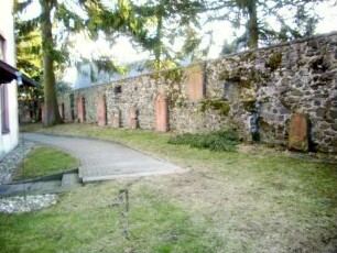 Kirchhof im Westen mit Kirchhofmauer (erhaltene Schießscharte in Brusthöhe - Wehrgang mit Steinbrüstung im 18 Jh abgetragen) sowie aufgestellten Grabsteinen aus dem Kirchhof