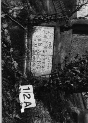 Grabstein 12 (Index: 100080863) Material: Sandstein mit weißer Marmorplatte im Sockel Zustand: Schriftplatte der Stele fehlt, Schriftplatte im Sockel gebrochen und auf dem Kopf, insgesamt stark verwittert, efeubewachsen Maße HxBxT: 135x57x16 cm Beschreibung: ädikulaartige Stele, Sockel, Rundbogenabschluß, eingetieftes Schriftfeld mit Rundbogen: Grabstein 12A