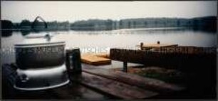 Blick auf einen See mit Enten, im Vordergrund Holzsitzbank und Campingkocher (Sonderthema: Guten Morgen)