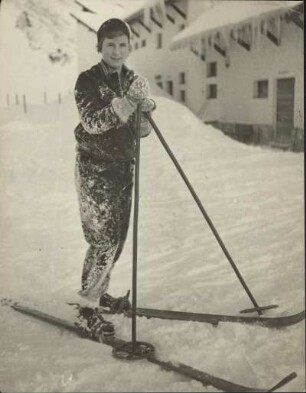 Eine Skifahrerin steht vor einer Skihütte. Sie scheint gestürzt zu sein - die Sachen sind voller Schnee