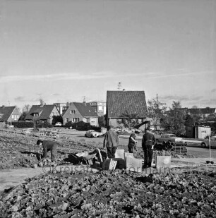 Katholische Kirchengemeinde St. Vicelin: Friedrich-Bölck-Straße: vor Einweihung: Herrichten der Außenanlagen: Arbeiter beim Anlegen des Plattenwegs zur Lorentzenstraße, Betonplatten, Schubkarre, Drainagerohre: Einfamilienhäuser an Lorenzenstraße, parkende Autos: im Hintergrund Theodor-Mommsen-Schule