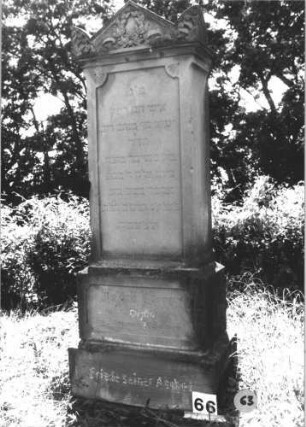 Grabstein 63 (Index: 99805298) Material: Sandstein Zustand: leicht verwittert Maße HxBxT: 182x69x41 cm Beschreibung: Stele, getreppter Sockel, Giebel mit stilisierten Eckakroteren, rechteckiges Schriftfeld eingetieft Symbolik: Blätterkranz im Giebel: Grabstein 63