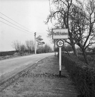 Landstraße mit Frostschäden: vorn rechts Hinweisschild: daneben Hecke und Bäume: links Strommast und Knick
