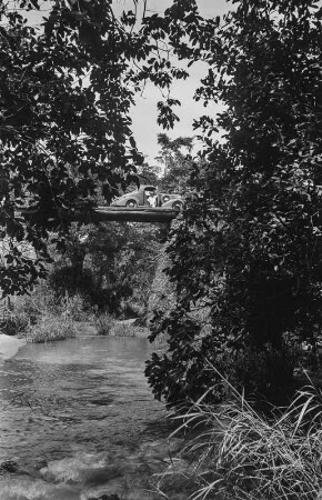Brücke : Schmale Brücke aus Baumstämmen, darauf Mann vor seinem Fahrzeug.