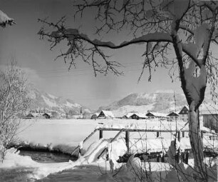 Winterbilder. Verschneite Brücke über einen Bach unweit eines Dorfes am Fuße eines Gebirges