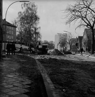 Mewesstraße: Blick Richtung Kurparkallee: links an Einmündung Reimer-Hansen-Straße Bagger: davor zwei Fußgänger: dahinter Kurparkhügel mit Bäumen und Kreishaus: hinten Mitte Hochhaus "Liebe": hinten rechts Wohnblocks