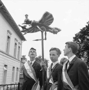 Kindervogelschießen: Umzug: Nahaufnahme: drei Schüler mit Schärpe, einer trägt Vogel, die beiden anderen schauen zu Vogel empor: rechts Teil von Blumenkrone: links Haus, 30. Juni 1964