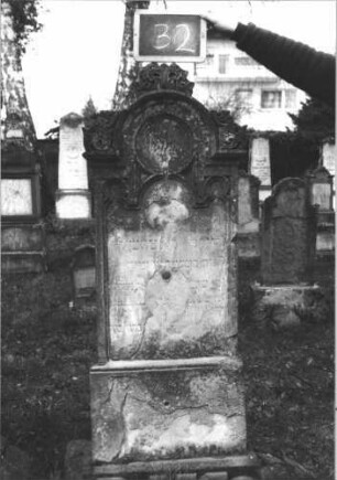 Grabstein 32 (Index: 100112294) Material: Sandstein Zustand: beschädigt und verwittert Maße HxBxT: 165x50x12 cm Beschreibung: Stele, getreppter Sockel, eingezogener Rundbogenabschluß mit Akroteren, Schriftfeld eingetieft mit Rundbogenabschluß Symbolik: Maßwerk: Grabstein 32